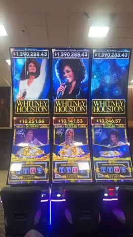 This video shows three "Whitney Houston" themed slot machines inside a casino, likely in Reno, United States of America. Each machine features a large image of Whitney Houston at the top and a digital display showing various jackpot amounts.

The prominent "PLATINUM" jackpot displays $1,390,288.4 across all three machines. Below this, "GRAND" jackpots are shown: $10,251.66 on the left machine, $10,141.53 on the middle, and $10,246.57 on the right. "MAJOR" jackpots are also displayed: $1,109.99 (left), $1,191.53 (middle), and $1,241.84 (right). A "MINOR" jackpot of $50.00 is visible on all three.

The lower part of each machine indicates options to "CHOOSE YOUR DENOMINATION," with choices including 1¢, 2¢, 5¢, 10¢, and 25¢. The machines are primarily gold and blue, with images of Whitney Houston in various poses. Black chairs with purple lighting are positioned in front of the machines. The general scene suggests a typical, well-lit casino environment.