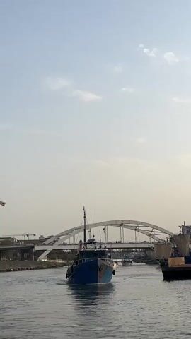 A blue and white fishing boat travels down a canal toward a white arched bridge. The water is calm and reflects the sky, which is mostly clear with a few wispy clouds. On the left bank of the canal, there is a construction crane and some buildings. On the right bank, there are other boats and structures. The sun appears to be low in the sky, casting a soft light on the scene.
