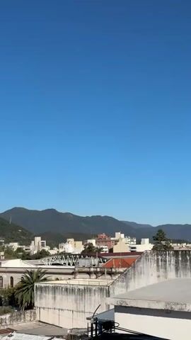 The video shows a scenic view of a town located at the base of mountains, under a clear blue sky. The camera slowly pans from right to left, revealing the cityscape, which includes buildings of varying heights and architectural styles, palm trees, and other greenery. In the background, a mountain range covered in lush vegetation stretches across the horizon. The scene appears calm and peaceful, with no visible human activity. The foreground consists of rooftops and architectural details of the buildings from which the video is shot.