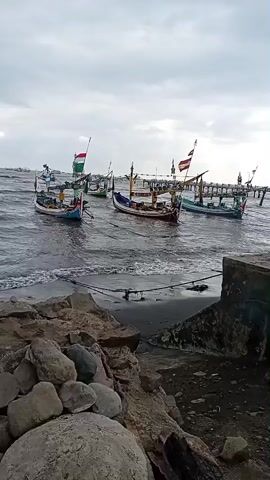The video shows a view of a rocky shore looking out onto the ocean under a cloudy sky. Several small fishing boats are visible on the water, some with flags. The waves are moderately choppy, lapping at the shore. In the foreground, large rocks and some debris are scattered on the beach. A pier or jetty can be seen in the distance on the right side of the frame. The overall scene is one of a working harbor or coastline, possibly before or after a fishing trip.