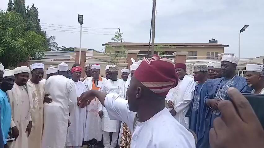 A group of men in traditional attire are gathered outdoors in front of a building and some trees. The men are mostly wearing white robes and caps, with some wearing elaborately patterned garments. The man in the foreground on the left is wearing a red and white striped cap and is gesturing with his right hand. Several people are looking towards him. The man in the foreground on the right is holding a camera or phone, partially obscuring his face. The environment appears to be a street or courtyard with a paved surface. The sky is overcast.