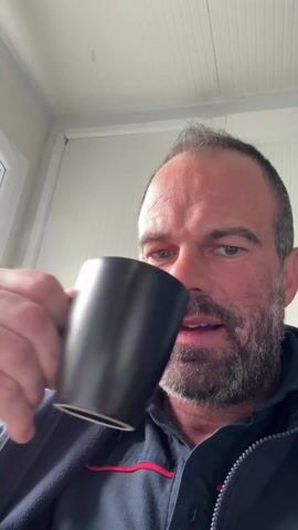 A middle-aged man with a beard is drinking from a black mug. He has graying hair, blue eyes, and is wearing a dark jacket with red stripes. After taking a sip, he opens his eyes wide and looks directly at the camera with a surprised expression. The background appears to be a room with white walls and a glimpse of a window frame.