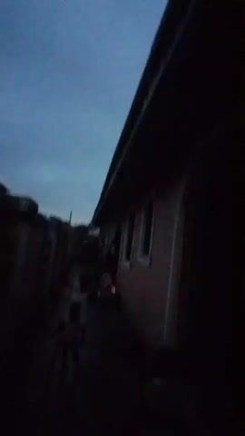 A dark video shows a person walking away from the camera down a narrow path between buildings. The person is blurry and appears to be a child. The sky is dark blue, suggesting dusk or dawn. There are buildings on either side of the path, one of which has windows. The ground is wet.