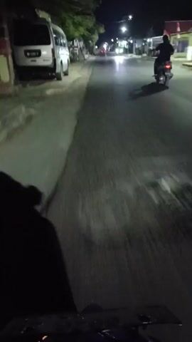The video shows a nighttime view from the perspective of someone riding a motorcycle. The road is paved and appears to be a street in a town or village, with buildings visible on either side. There are other motorcycles on the road, some with their headlights on, creating streaks of light. The rider is moving at a moderate speed, as indicated by the motion blur. The scene is dimly lit by streetlights and the headlights of the vehicles. Trees and foliage are also visible along the sides of the road. The overall impression is of a late-night journey through a quiet, semi-urban environment.