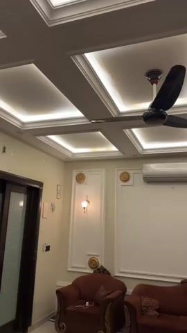 A shot of the interior of a room, focusing on the ceiling. The ceiling features a coffered design with recessed lighting. A ceiling fan is visible in the upper right of the frame. A sliding glass door with dark wood frames is on the left side of the room. Two plush armchairs, one on the left and one on the right side of the frame, are upholstered in a dark reddish-brown fabric. A couch is partially visible on the right. The walls are a light, neutral color. There is a decorative sconce light on the wall between the door and the armchairs.