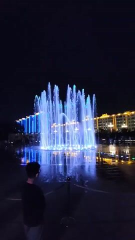 A person stands on a wet surface, looking at a large fountain display. The fountain sprays water in various patterns and is illuminated with colorful lights, primarily blue and green. In the background, buildings are visible with lights and decorative elements. The scene takes place at night.
