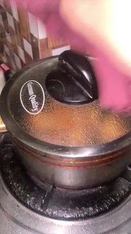 A person removes a lid from a dark-colored pot on a gas stove with a purple cloth. Inside the pot, chunks of browned chicken are frying in oil. The stove's burners are lit, and the pot is sitting on a black, stained stovetop. The background includes a tiled wall with a brown and white pattern.