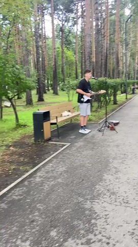 A young man stands on a paved walkway in a park, playing an electric guitar and singing into a microphone. He is wearing a black t-shirt, grey shorts, and white sneakers. A bench with a trash can next to it is to his left. There is a guitar case and an amplifier on the ground near him. Tall trees with green foliage surround the area.