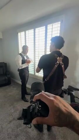 The video shows a man in a white dress shirt, black vest, and black pants adjusting his cufflinks in front of a window with white shutters. He is being filmed by another person holding a camera. The room is carpeted and has a black recliner chair on the left. The person filming is wearing a black shirt with a leather camera harness. The lighting is natural, coming from the window.