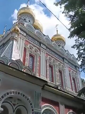 The video shows the exterior of the Russian Orthodox Church of the Nativity, commonly known as the Shipka Monastery, located in Shipka, Bulgaria. The building is characterized by its vibrant red walls, intricate white and grey decorative trim, and numerous golden domes topped with crosses. A tall, white bell tower with similar decorative elements stands to the left of the main structure. The camera pans across the facade of the church, highlighting its architectural details. The sky above is blue with scattered white clouds, suggesting a clear, possibly sunny day. Lush green leaves from a nearby tree are visible in the upper left corner, framing the scene. The overall impression is one of grandeur and religious significance, showcasing a well-maintained and historically important religious site. There is no visible text in the video. The time of day appears to be daytime, based on the lighting. The scene is static, with no people or immediate activity shown, focusing solely on the impressive architecture of the monastery.