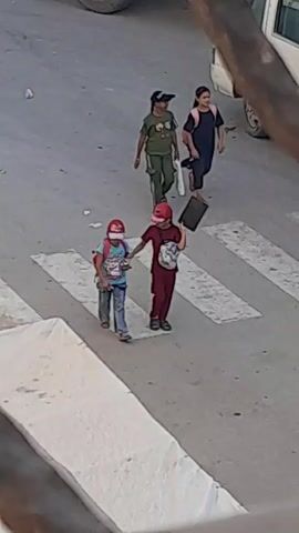 Four children are walking across a pedestrian crossing. Two children in the foreground are walking side-by-side. The child on the left is wearing a blue t-shirt, blue jeans, and a pink baseball cap. The child on the right is wearing a maroon tracksuit and a red baseball cap, carrying a black bag on their shoulder and holding a black tablet. Behind them, two other children are also walking. The child on the left of this pair is wearing a green t-shirt with a cartoon character, green pants, and a black baseball cap, carrying a white bag. The child on the right of this pair is wearing a dark blue t-shirt, dark pants, and a pink backpack. The ground is asphalt with white pedestrian crossing markings. A vehicle is partially visible in the upper right corner of the frame. The lighting suggests it is daytime.