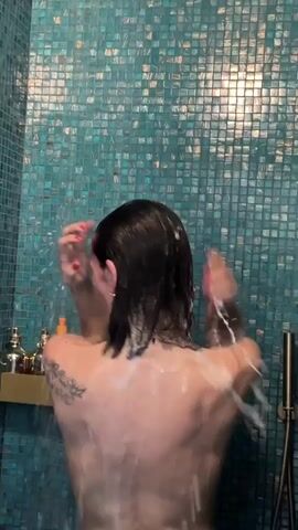 A person, from the back, is showering in a shower stall tiled with small teal-colored square tiles. A small gold-colored shelf with bottles is visible on the left wall. The person has dark, short hair, fair skin, and a tattoo on their left shoulder. They are applying soap to their hair and body under the running water. Their nails are painted red. The camera angle is stationary and focuses on the individual in the shower.