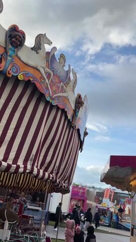The video shows a scene at an outdoor fair or amusement park. The sky is overcast with blue patches. The video starts with a close-up of a carousel structure with horses and angels on it, with a red and white striped awning. A Ferris wheel with a sign reading "Kermis Maaseik" and "Bon Voyage" is visible. People walk around the fair. Next, the Ferris wheel is shown in its entirety, followed by a tall drop tower and a sign for a "Caribbean Beach Party." The video then shows a spinning ride with people on it. The Ferris wheel is shown again before the video ends.