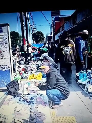 The video appears to be a series of short clips taken at an outdoor market. The first clip shows a bustling market scene with many people browsing stalls laden with various items, including clothing, shoes, and what appear to be trinkets or small decorative objects. The stalls are set up on the ground and along a sidewalk. Graffiti is visible on a wall in the background, and there are electrical wires overhead. The second, third, and fourth clips provide closer views of the merchandise, focusing primarily on shoes and various metal objects displayed on tables. The overall impression is of a vibrant, somewhat chaotic street market environment.