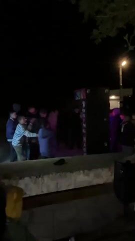 The video captures a nighttime scene in Iganovo, Bulgaria, where a group of approximately 10-15 young men and boys are gathered around a large, black arcade-style boxing machine. The machine, which has "BOXING MACHINE" written vertically in white letters on its side, is illuminated by colorful lights on its top and front edge, contrasting with the dark surroundings.

A young man wearing a white puffer jacket steps forward and delivers a forceful punch to the boxing machine. The impact is audible. The other individuals in the group watch intently, their attention focused on the action. Some are leaning in closer, while others stand back, observing the participant. A street light pole is visible in the background, and sparse tree foliage can be seen at the top of the frame. A low concrete wall is in the immediate foreground. The casual attire of the participants and the setting suggest a moment of leisure and entertainment in a public outdoor space.