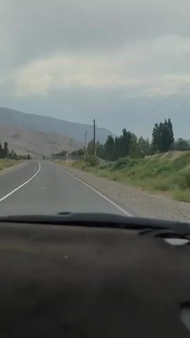 The video shows a view from inside a car, looking out at a two-lane road stretching into the distance. The road is paved and has white lane markings. To the left of the road, there are mountains with dry, barren slopes. To the right, there is a grassy area with scattered trees and utility poles. The sky is overcast with clouds. The car's dashboard is visible in the foreground. The video appears to be shot while the car is in motion, driving along the road. There are no other vehicles or people clearly visible in the immediate vicinity.