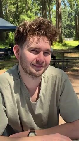 The video shows a close-up shot of a young man with curly brown hair, wearing a light green t-shirt. He is sitting at a table outdoors, with a blurred background of trees and a picnic table. The man is looking at the camera and smiling slightly. The lighting suggests it is daytime, likely a sunny day, as there are shadows cast on the man's face and shirt. He is wearing a watch on his left wrist. The video appears to be a candid shot, possibly taken by someone seated across from him at the table.
