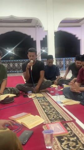 The video shows five young men sitting on a red carpet in a room with arched openings and white walls. The man on the far left is speaking into a microphone, holding an open book in front of him. He is wearing a black t-shirt and dark pants. To his right, three other men are seated, also with open books in front of them. They are wearing dark shirts and pants. The man closest to the microphone is wearing a blue shirt. The next man is wearing a dark olive green shirt. The furthest man on the right is wearing a maroon shirt and a black skullcap. Another person's arm is visible on the far left, also holding a book. There are several clear plastic cups and what appear to be more open books scattered on the carpet. The overall lighting suggests it is evening or indoors. The men appear to be engaged in some form of recitation or lesson.