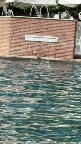 This is a close-up shot of a swimming pool with clear turquoise water. In the background, a brick wall with a rectangular white cutout is visible. Water flows from the cutout in a thin sheet, and two small fountains spray water upwards from the top of the brick structure. The scene is illuminated by natural sunlight, casting reflections on the water's surface. Umbrellas and a glimpse of white chairs are visible behind the brick wall, suggesting an outdoor setting like a resort or patio. There are no people clearly visible in the frame.