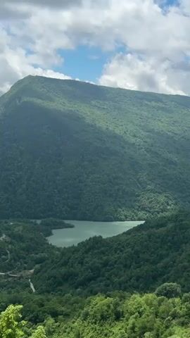 The video shows a drone flight over a lush green mountainous landscape. In the foreground, dense green trees are visible, with their leaves appearing vibrant and healthy. As the drone ascends, a tranquil lake comes into view, nestled within the valley. The lake has a serene, almost mirror-like surface, reflecting the surrounding greenery. Further into the distance, a large, majestic mountain dominates the horizon, its slopes covered in a thick carpet of trees. The sky above is a mix of blue and white, with fluffy clouds scattered across it, suggesting fair weather. The overall impression is one of natural beauty and peaceful serenity, with the drone's movement allowing for a panoramic exploration of the picturesque scenery.