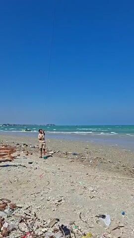 A person is standing on a beach, holding a kite string. The beach is littered with trash. The ocean is in the background, with waves breaking on the shore. The sky is clear and blue. There are boats visible in the distance. The person is wearing a light-colored shirt and shorts.
