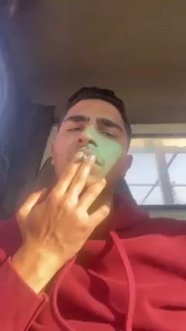 A young man wearing a red hoodie is sitting in what appears to be a vehicle. He is smoking, as smoke is visible coming from his mouth. His face is partially illuminated by sunlight, creating a greenish hue on part of his face. The interior of the vehicle is visible in the background.