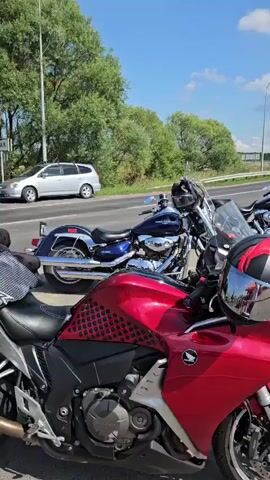 A group of motorcycles are parked on the side of a road. A red motorcycle is in the foreground, with a helmet resting on its handlebars. Several other motorcycles, including a blue cruiser and a yellow sportbike, are parked behind it. In the background, a white car and a white SUV are visible on the road, along with a road sign indicating directions to Kemerovo and Leninsk-Kuznetsky. The sky is blue with scattered clouds, and trees line the side of the road. The scene appears to be a rest stop or meeting point for motorcycle riders.