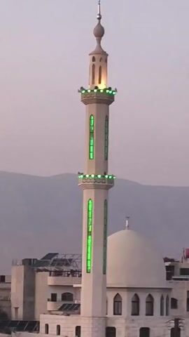 A medium, full shot captures a mosque's minaret and dome. The minaret, positioned prominently in the center, is illuminated with green lights along its length and a soft golden glow emanating from its top. A silver crescent moon adorns the dome of the mosque. The background features rooftops of surrounding buildings and a mountain range in the distance. The sky is a muted, light color, suggesting either early morning or late evening. The camera is static, maintaining a fixed viewpoint throughout the duration of the shot.