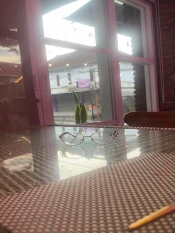 The video features a close-up shot of a pair of rose gold-colored eyeglasses resting on a clear glass table. The table has a textured, possibly wicker or rattan, base visible underneath the glass surface. Through a large window with light pink frames behind the table, an outdoor scene is visible, including houses, a street, and tall green snake plants. Small string lights are reflected on the glass tabletop and the lenses of the eyeglasses, suggesting a warm and ambient lighting. The camera slowly zooms in on the eyeglasses, highlighting their intricate design and the reflections of the lights. The outdoor background becomes progressively more blurred as the zoom intensifies, bringing the eyeglasses into sharper focus.