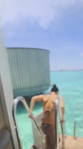 The video shows a woman diving into clear turquoise water from a wooden deck. She is seen from the back, briefly raising her arms before jumping in. The camera then follows her underwater for a moment. After that, the video shows the woman walking down a wooden boardwalk between buildings towards the sea, carrying a yellow bag. The sky is blue with some clouds.