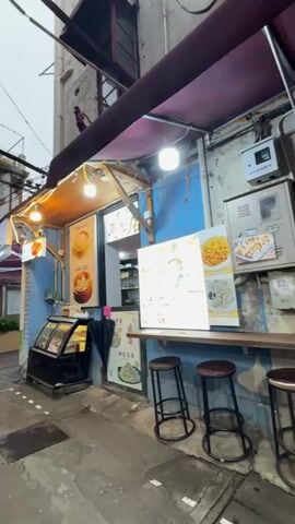 The video shows a small, quaint food stall tucked away in a narrow alleyway. The stall is painted a vibrant blue and features a wooden awning adorned with string lights. Posters displaying pictures of various dishes, including dumplings and noodles, are prominently displayed. A black display case filled with treats sits near the entrance, and a small table with two stools offers a place for customers to sit and eat. The alleyway appears somewhat wet, and buildings can be seen rising on either side.
