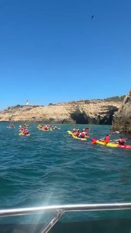 The video shows a group of people in kayaks on the ocean near a rocky coastline under a clear blue sky. The kayaks are various colors, including red and yellow, and many of the people are wearing red life vests. They appear to be on a guided tour or excursion, with several kayaks clustered together. In the background, there is a lighthouse on a cliff overlooking the sea. The water is blue and appears calm. The perspective of the video suggests it was filmed from a boat.