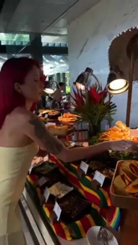 A woman with red hair and a light yellow strapless dress is standing at a food buffet, filling a small white plate with items from the buffet. The buffet is decorated with a colorful Mexican-style striped cloth and has various dishes, including tortilla chips, beans, and other Mexican food items. Above the buffet are heat lamps and a decorative sombrero. Other people are visible in the background, as well as large windows showing outdoor greenery.