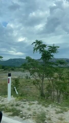 The video captures a scenic view from inside a moving vehicle. The camera is positioned to show the side mirror, reflecting a portion of the interior and the driver's side. The landscape outside is characterized by rolling hills and mountains in the distance, under a cloudy sky. The terrain in the foreground appears to be a mix of dry grass, sparse vegetation, and rocky patches. A white pole with a red top is visible on the left side of the frame. The overall impression is that of a journey through a rural or semi-arid environment.