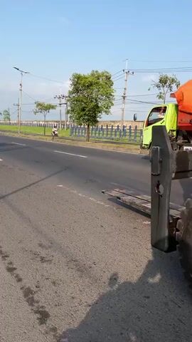 The video shows a yellow Caterpillar forklift with the number 100 visible on its side, driving on an asphalt road. A person wearing an orange safety vest and a grey shirt is operating the forklift. The forklift has large black tires and a metal frame with a roof. The environment appears to be outdoors with a clear blue sky and some trees in the background. The forklift is moving forward, and the camera is positioned to the side of the vehicle.
