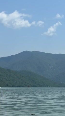 A calm body of water stretches across the foreground, reflecting the sky and distant mountains. The water is a greenish-blue color and appears rippled by a light breeze. In the middle ground, two boats are visible on the water. The boat on the left is a small speed boat with a person or two visible onboard, moving at a moderate speed creating a wake. The boat on the right is a smaller, red-topped vessel, seemingly stationary or moving very slowly. Behind the water, a series of densely forested mountains rise, their slopes covered in lush green vegetation. The largest mountain is in the center background, with a rounded peak. The sky above is a clear blue, dotted with a few fluffy white clouds. The overall scene depicts a serene natural landscape with a touch of human activity on the water.
