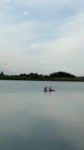 Two people paddle kayaks on a calm lake with trees and houses on the far bank under a cloudy sky.