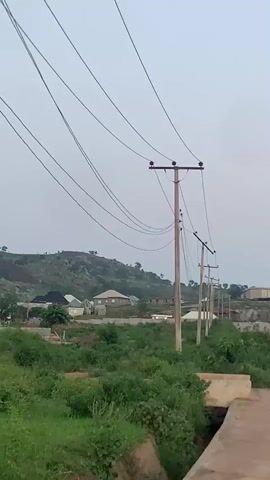 The video pans from left to right, showing a road in what appears to be a rural or developing area. On the left side of the road, there are several utility poles with power lines stretching across the frame. Beyond the poles, there are some houses and vegetation. As the video pans to the right, the road curves and continues into the distance. In the background, there are hills covered with rocks and vegetation. A few buildings are visible, some with green roofs. The sky is overcast. There are no people clearly visible in the video, and no specific actions or interactions are depicted. The environment appears to be outdoors, possibly in the morning or evening due to the lighting.