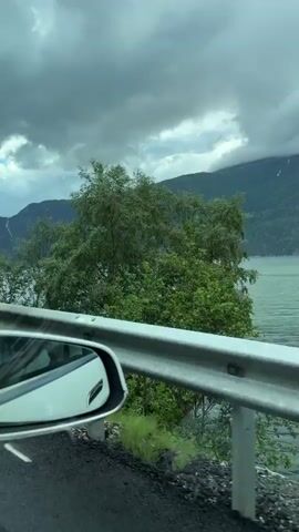 The video shows a view from a car window, looking out at a large body of water, possibly a fjord or lake, with steep, tree-covered mountains in the background. The sky is cloudy and grey. A small boat is visible on the water in the distance. A white side-view mirror of the car is in the lower left corner of the frame, reflecting the scenery. A metal guardrail runs along the side of the road in the foreground. The road itself is visible, with a white line marking the lane. Green grass and some rocks are present on the roadside. The overall atmosphere is overcast and possibly cool.