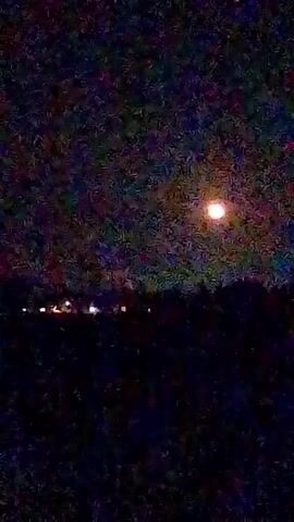 A full moon is visible in the night sky, casting a dim glow over a dark landscape. The moon appears as a bright, circular object with a reddish-orange hue, positioned in the upper right quadrant of the frame. The sky is speckled with what appears to be stars or noise, creating a textured effect. In the middle ground, a faint line of lights suggests the presence of buildings or structures, possibly illuminated by the moonlight or their own internal lighting. The foreground is entirely dark, obscuring any details of the ground. The overall scene is one of a quiet, nighttime setting, with the moon as the prominent celestial feature.