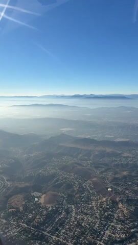 This is an aerial view of a landscape. In the foreground, there are hills covered in vegetation and houses. In the midground, there are more hills and a layer of fog or clouds. In the background, there are mountains. The sky is clear and blue. The sun is shining. The video is taken from an airplane or helicopter.