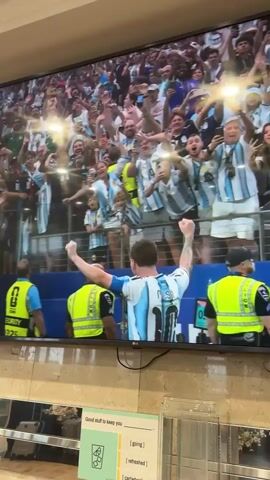 A large television screen displays a soccer match between Argentina and Austria, with Argentina leading 2-0 in the final. The screen also shows fans in the background wearing Argentina jerseys and waving flags. In the foreground, two children, also in Argentina jerseys, hold up signs. One sign reads "HAPPY EARLY BIRTHDAY MESSI!". The other sign displays "GOAL KING THE CU" with a crown graphic. The scoreline is visible at the bottom of the screen, indicating "GROUP J ARGENTINA 2 AUSTRIA 0 FINAL". The setting appears to be indoors, possibly a public space or a room with a hotel-like aesthetic, as suggested by the tiled wall and a brochure on a table below the TV that reads "Good stuff to keep you [going] [refreshed]". The lighting suggests it is daytime. The atmosphere appears celebratory and enthusiastic, with the focus on Lionel Messi, evident from the birthday wishes.