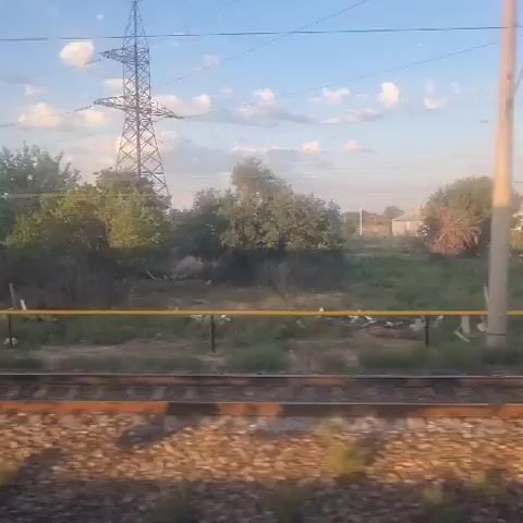 The video shows a view from a moving train, looking out at a rural landscape. In the foreground, train tracks are visible, with ballast and ties. Beyond the tracks, there is a field of green grass and some scattered trees. A yellow pipe runs horizontally across the middle ground, supported by short posts. In the background, there are a few small buildings and a large power line tower. The sky is blue with scattered white clouds. The lighting suggests it is daytime, possibly late afternoon or early morning due to the warm tones. The scene is static except for the implied movement of the train. There are no people or animals visible.