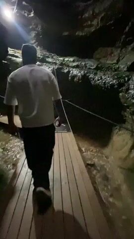A person walks on a wooden boardwalk through a cave. The boardwalk is elevated above murky water. The cave walls are rough and dark, with some areas illuminated by artificial lights. The person is wearing a white t-shirt and dark pants. They are walking away from the camera, towards the illuminated opening of the cave.
