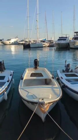 The video shows a marina with several boats docked. In the foreground, there are three small white motorboats. The boat in the center has a wooden pole sticking up from its bow. Behind these boats, larger yachts and sailboats are moored. The water is calm and reflects the sky and the boats. The sky is clear and blue, indicating a sunny day. The overall scene is peaceful and serene.