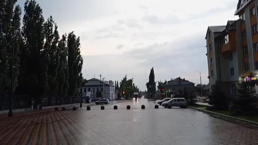 The video opens with a wide shot of a street on a cloudy day. On the left, a line of tall, dark green trees stands in front of a building. A wet, brick-paved walkway is visible in the foreground with several dark, spherical bollards spaced evenly across it. The street itself is wet and reflects the overcast sky. Cars are visible on the street, and some appear to be moving. Traffic lights are red in the distance. On the right side of the street, there are several buildings, including a multi-story apartment building with balconies. The sky is filled with gray, scattered clouds, with a lighter hue visible near the horizon, suggesting either early morning or late evening. The video continues with slightly varying shots of the same scene, with some close-ups of the clouds and the general streetscape.