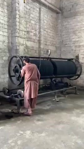 A man is shown from behind, operating a large, industrial machine in a room with exposed brick walls. The machine consists of two large, cylindrical drums with a series of ridges, and a smaller wheel at the base that appears to be a drive mechanism. The man, dressed in a long, peach-colored shirt and matching pants, appears to be adjusting something on the machine. The room seems industrial, with a concrete floor and minimal decor. There's a blue container visible in the background on the left side of the frame. The scene is static with no camera movement.