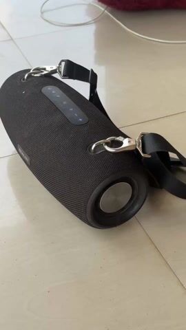 A black, cylindrical Bluetooth speaker with a black shoulder strap rests on a beige tiled floor. The speaker has silver hardware attaching the strap. There is a row of buttons on the top of the speaker. The ends of the speaker are round with a circular silver center. In the background, a red, fuzzy object is partially visible along with a white cable.