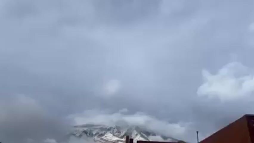 The video shows a snow-covered mountain partially obscured by clouds under an overcast sky. A rooftop with some structures is visible in the foreground. The general environment appears to be a residential or built-up area, given the presence of the rooftop and structures. The camera angle is slightly upward, focusing on the mountain and sky.