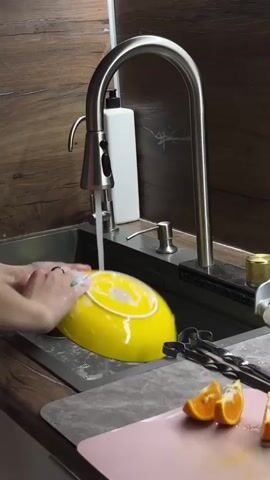 A person's hands are washing a yellow and white bowl under running water from a kitchen faucet. The sink is made of stainless steel. To the right of the sink, on the countertop, are two pieces of sliced orange on a pink cutting board, and a pair of tongs. Behind the faucet, there is a white bottle of soap and a silver dispenser. The wall behind the sink is made of wood-paneled material.